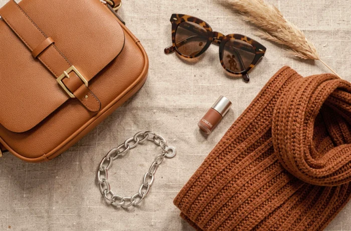 Flat-lay d'accessoires de mode tendance 2024 : sac en cuir caramel, lunettes écaille, bracelet chaîne et écharpe en maille rouille